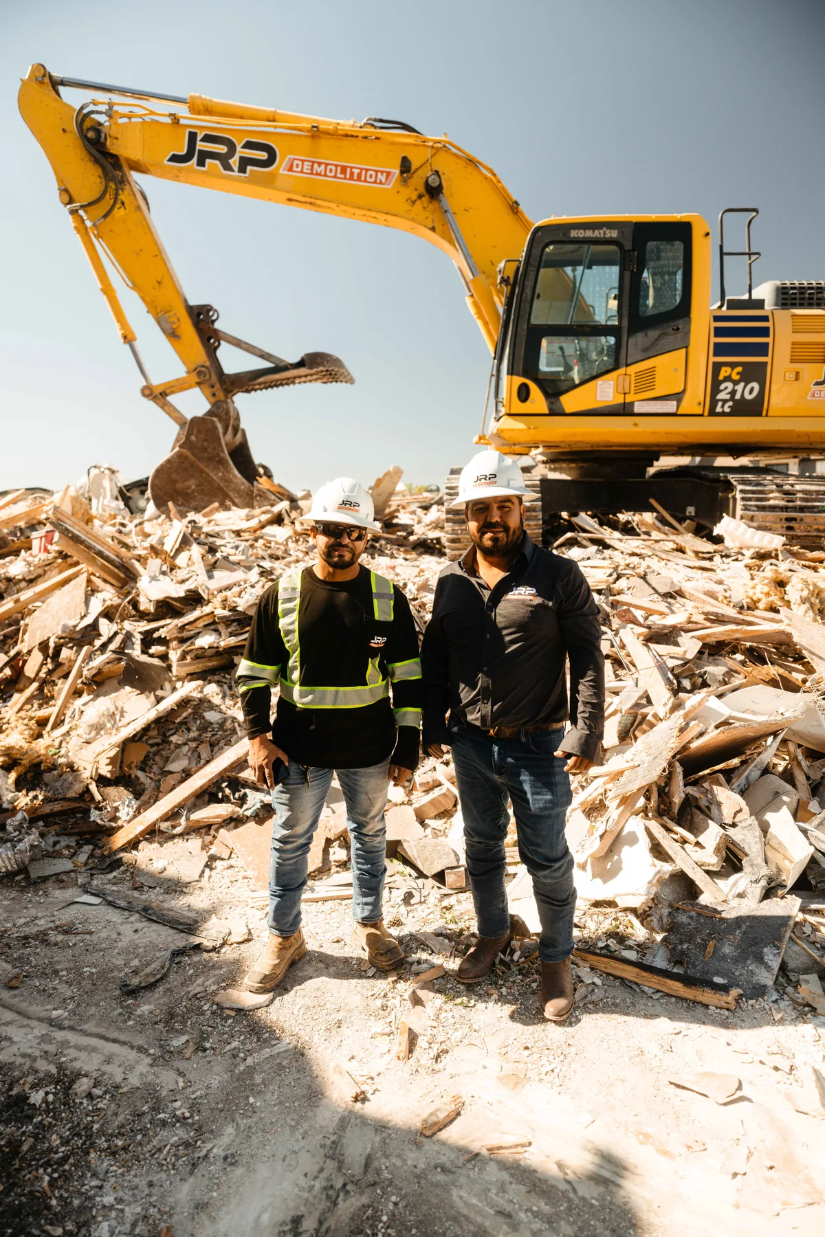Two JRP Demolition crew members in safety gear standing in front of a hydraulic excavator at a residential demolition job site in Dallas